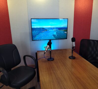 Estúdio de podcast com duas cadeiras de escritório posicionadas em frente a uma mesa de madeira. Sobre a mesa, há dois microfones fixados em suportes de base. Na parede ao fundo, revestida com painéis acústicos em vermelho e bege, há uma televisão ligada exibindo uma imagem aérea de uma paisagem com mar e barcos.
