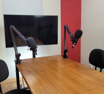 Estúdio de gravação de podcast com duas cadeiras posicionadas em frente a uma mesa de madeira. Dois microfones com suportes articulados estão fixados na mesa, voltados um para o outro. Ao fundo, há uma televisão ou monitor desligado montado na parede, que é revestida com painéis acústicos em cores alternadas de vermelho, bege e cinza-claro.