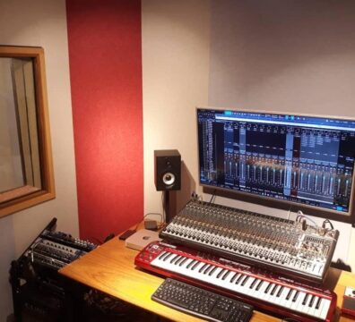 Imagem de uma sala técnica de estúdio de áudio com equipamentos de produção musical. Sobre a mesa de madeira há um teclado controlador vermelho, uma mesa de som analógica, um teclado de computador e um monitor grande exibindo software de edição e mixagem de áudio. Ao lado do monitor há dois monitores de áudio (caixas de som) e um microfone. À esquerda, uma janela de vidro permite ver a cabine de gravação ao fundo.